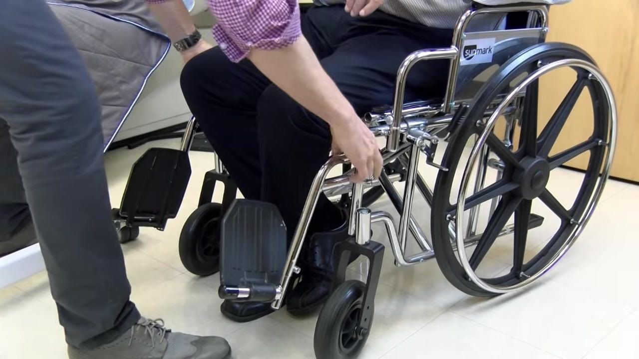 A caregiver locks the wheelchair brakes and prepares to swing away or remove the leg rests while the patient remains seated.
