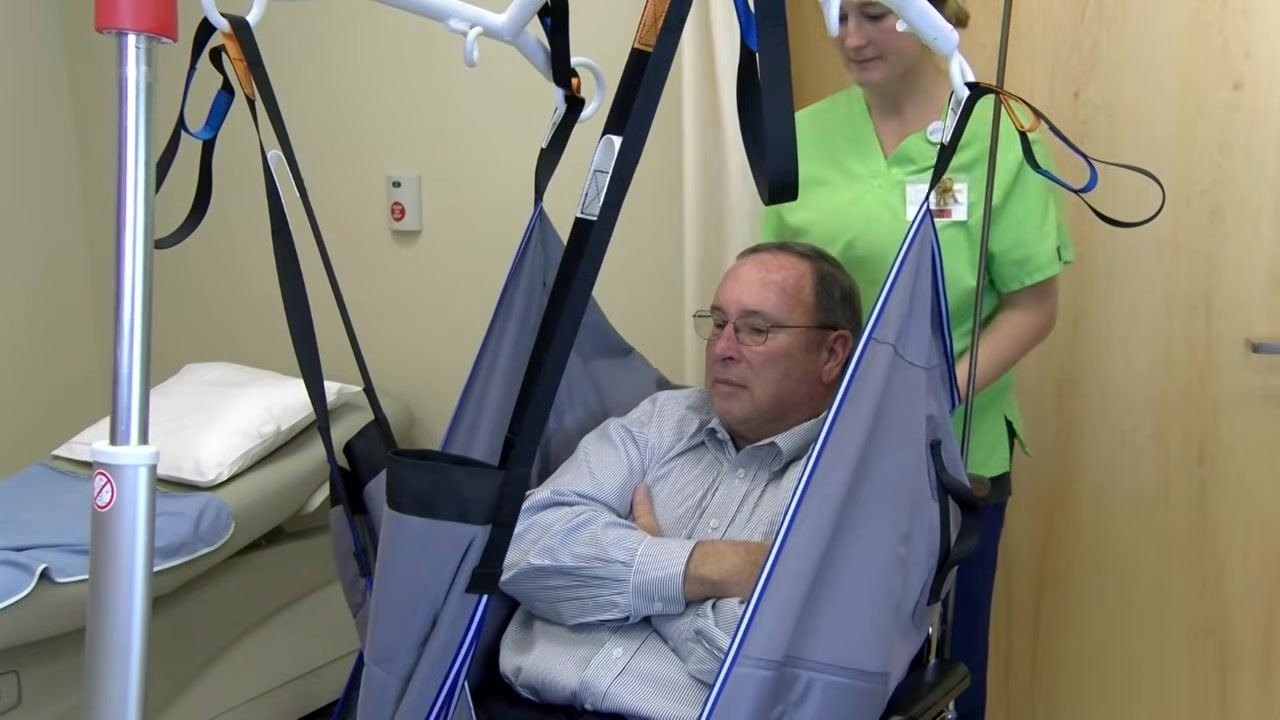 The patient sits in the sling with arms crossed inside it, while a healthcare worker stands behind, preparing for the lift.