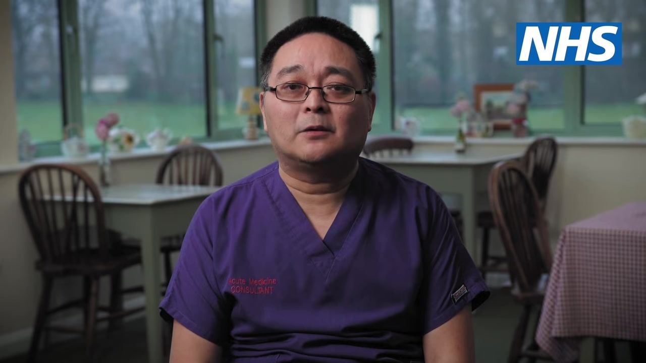 A healthcare professional in purple scrubs labeled "Acute Medicine Consultant" speaking in a calm indoor setting with NHS branding visible.