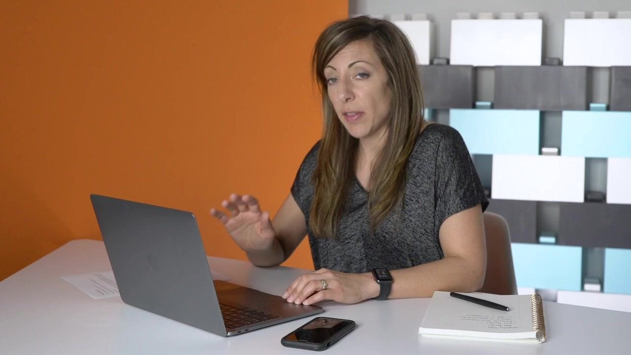 A person sitting at a desk with a laptop, smartphone, and notebook, gesturing as if explaining or demonstrating something. The environment is a modern office with an orange accent wall and a geometric patterned background.