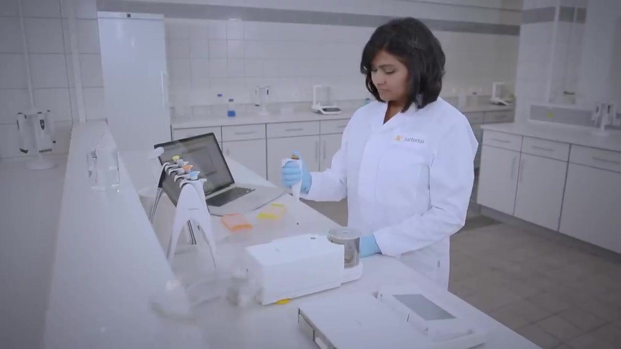 A laboratory technician in a white lab coat and blue gloves pre-wetting a pipette tip at a calibration station. The workspace includes a balance, pipette stand, disposable tip boxes, and a laptop.