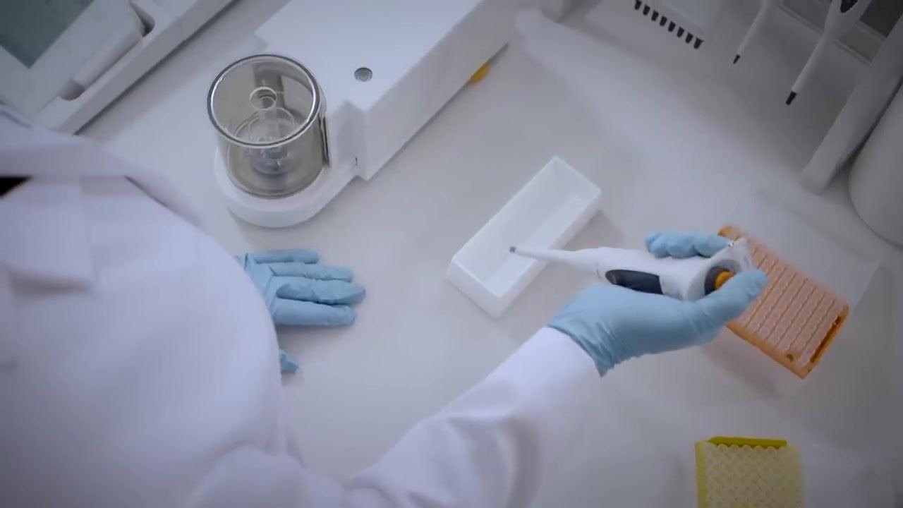 Overhead view of a gloved technician holding a pipette at a 45-degree angle, dispensing liquid into a weighing vessel on a balance. Pipette tip boxes and calibration equipment are visible.