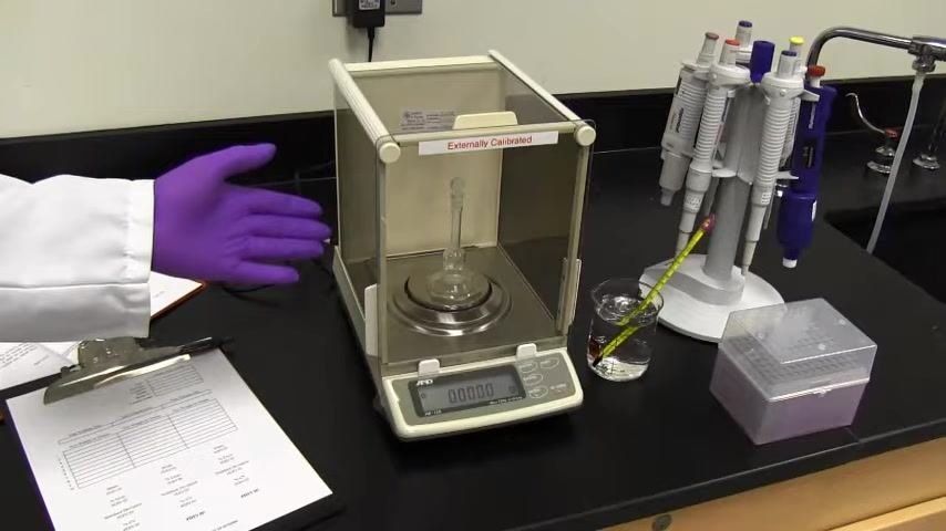 Laboratory setup showing the SOP document, documentation form on a clipboard, analytical balance with volumetric flask, beaker with thermometer, pipette stand with multiple pipettes, box of pipette tips, and a gloved hand indicating readiness.