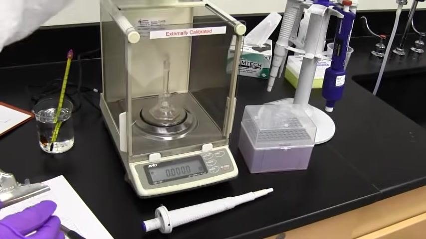 Analytical balance with a flask inside, display reading 0.0000 g, with the calibration worksheet nearby.