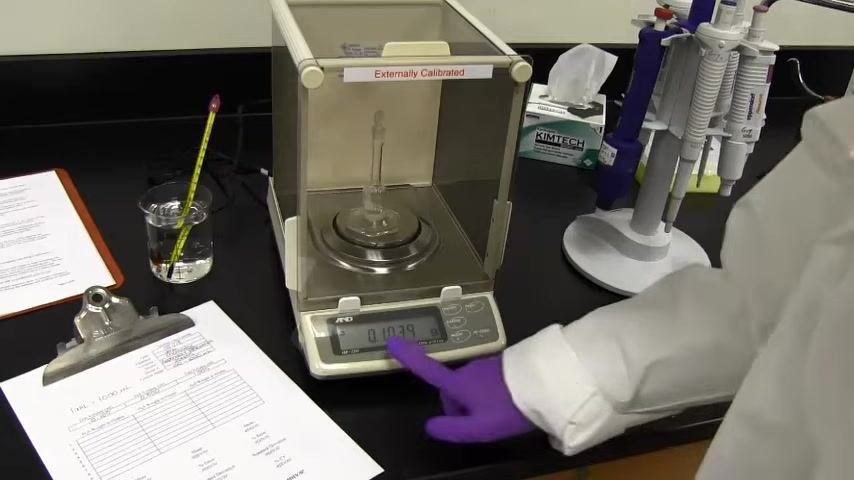 Balance display showing 0.1029 g, with the flask inside and worksheet visible; a gloved hand points to the reading.
