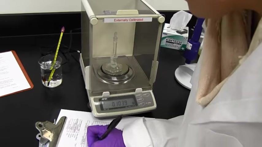A gloved hand records a weight (0.1039 g) on the calibration worksheet. The analytical balance displays 0.1039 g, with the flask on the balance, a thermometer in a beaker, Kimtech wipes, a pipette stand, and other lab items visible.