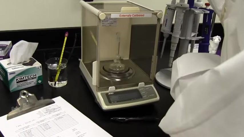 Analytical balance with a flask inside, labeled "Externally Calibrated." Kimtech wipes, thermometer in a beaker, pipette stand, calibration worksheet, and pen are visible, with a person in a lab coat present.