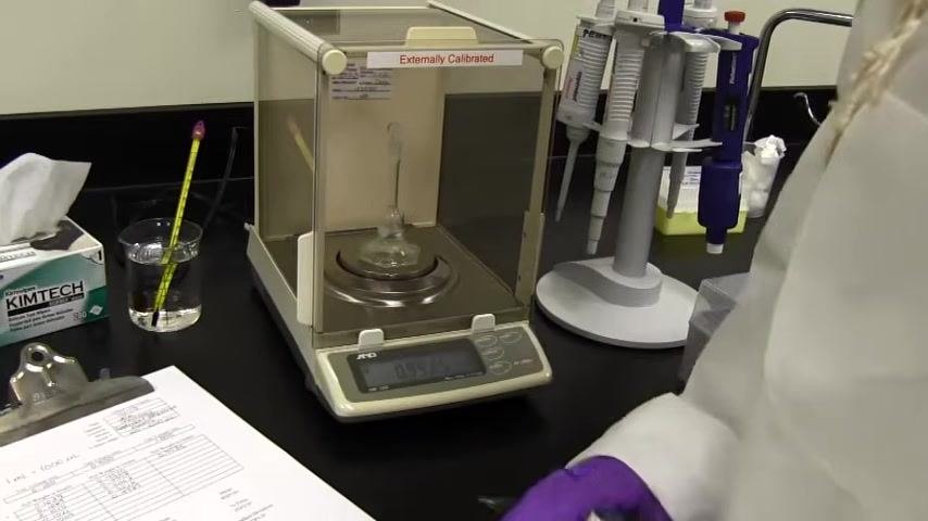 Analytical balance with a flask inside, display shows a weight (0.9975 g); a person in a lab coat and purple gloves is recording data, with the calibration worksheet, pen, thermometer, Kimtech wipes, and pipette stand visible.