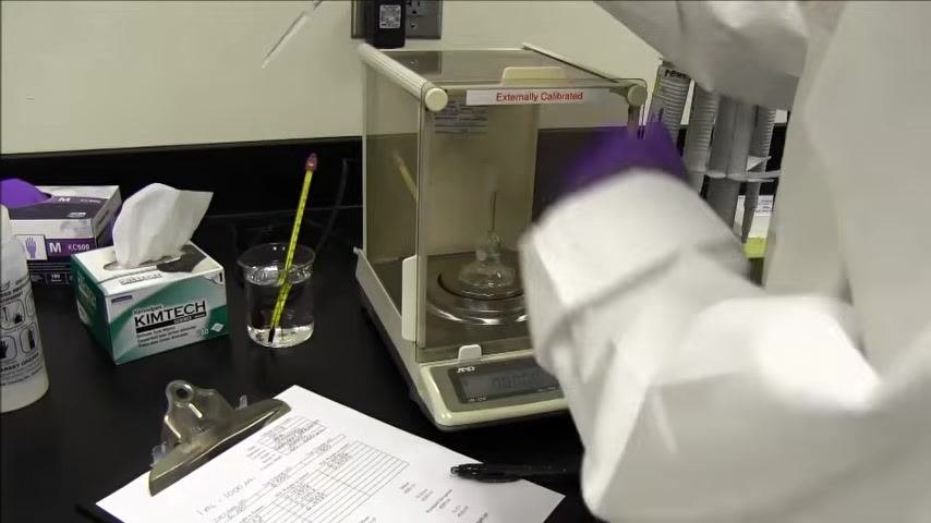 Person in a lab coat and purple gloves holds a pipette and pen, with the analytical balance, calibration worksheet, and pipette stand visible.