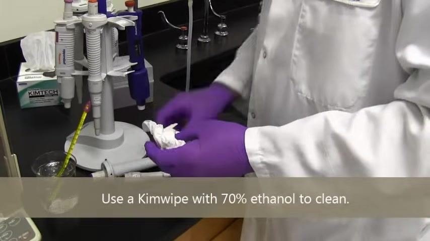 A person in a lab coat and purple gloves holds a pipette and a bottle of 70% ethanol, preparing to clean the pipette.