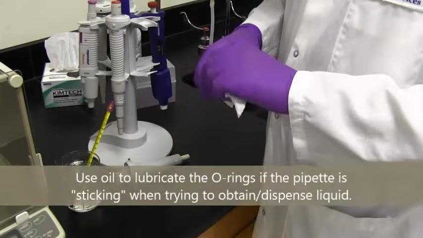 A person in a lab coat and purple gloves uses a Kimwipe with 70% ethanol to clean a pipette, with pipettes and lab equipment visible on the bench.
