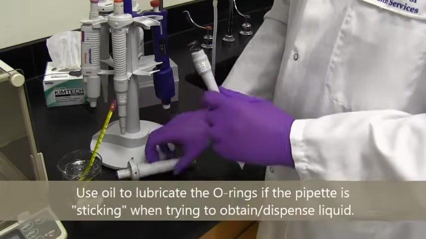 A person in a lab coat and purple gloves inspects and handles a reassembled pipette, with pipettes and lab equipment on the bench.
