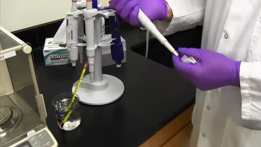 A person in a lab coat and purple gloves holds a pipette near a balance, a beaker with a thermometer, a pipette stand, and a box of Kimtech wipes.