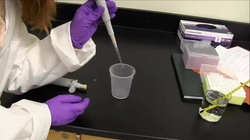 Gloved hand holding the pipette with the tip submerged in a beaker of water and a thermometer; on-screen text reads: "Once the tip is submerged, release your thumb to draw the water in."