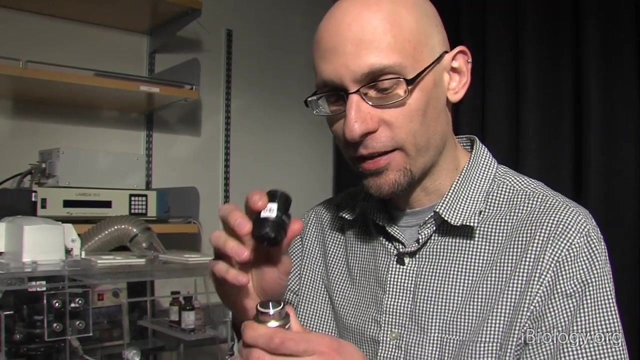 A person holds a microscope objective and an eyepiece, preparing to use the eyepiece as a magnifying glass to inspect the objective lens, with laboratory equipment and shelves visible in the background.