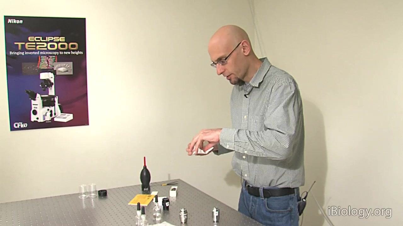 A person holds a cotton swab and prepares to clean a microscope objective with a recessed front element at a laboratory table, with cleaning supplies and objectives visible.