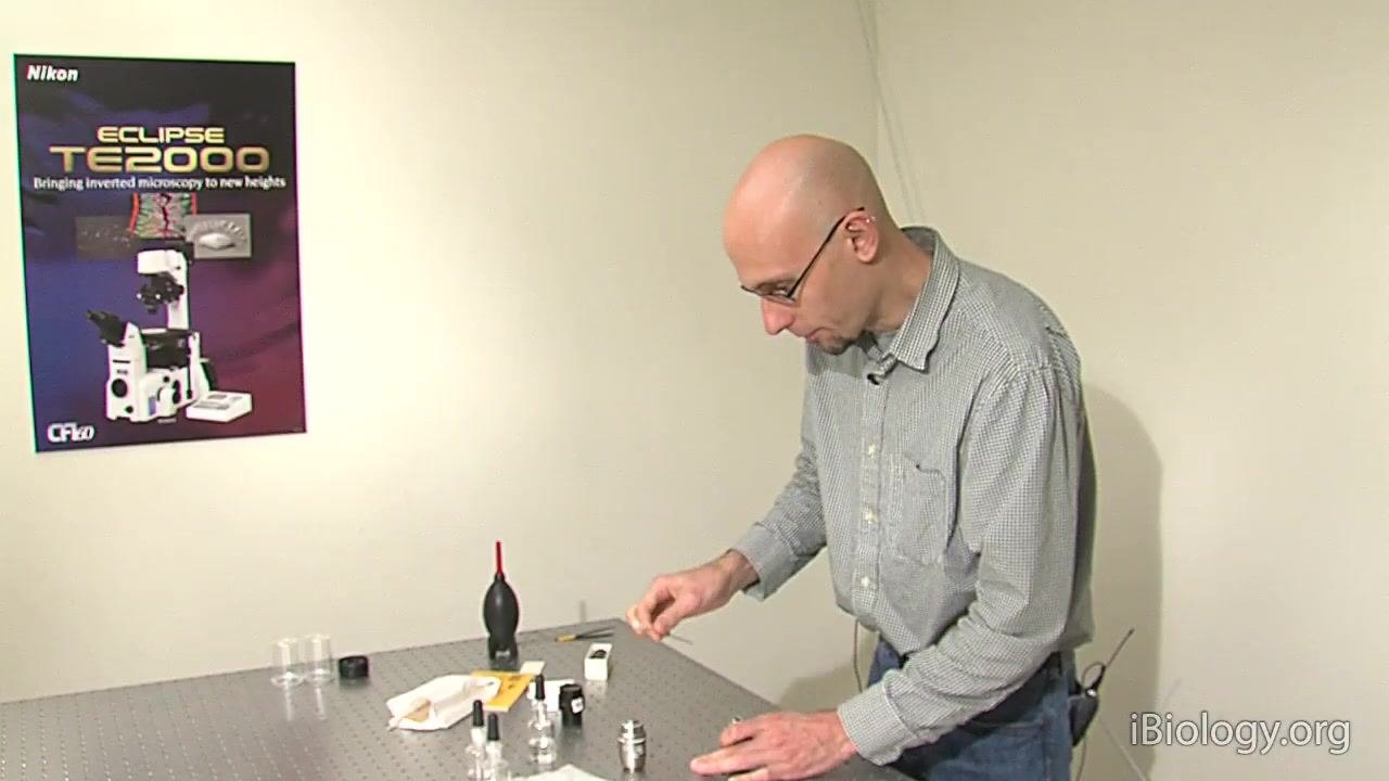 A person uses a cotton swab to clean a microscope objective, preparing to repeat the process with methanol, with cleaning supplies and objectives visible on the table.
