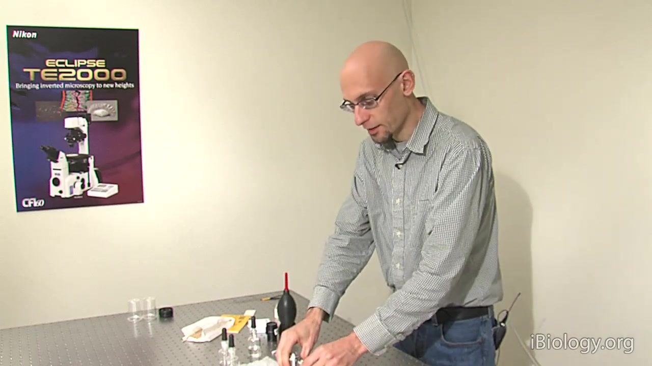 A person explains the final rinse steps for cleaning objectives, with cleaning supplies and objectives visible on the table.