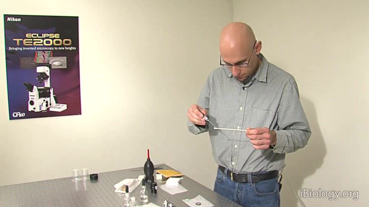 A person stands at the workstation, holding a cotton swab and preparing to moisten it with solvent, with cleaning supplies, lens paper, filters, and a Nikon ECLIPSE TE2000 poster visible.