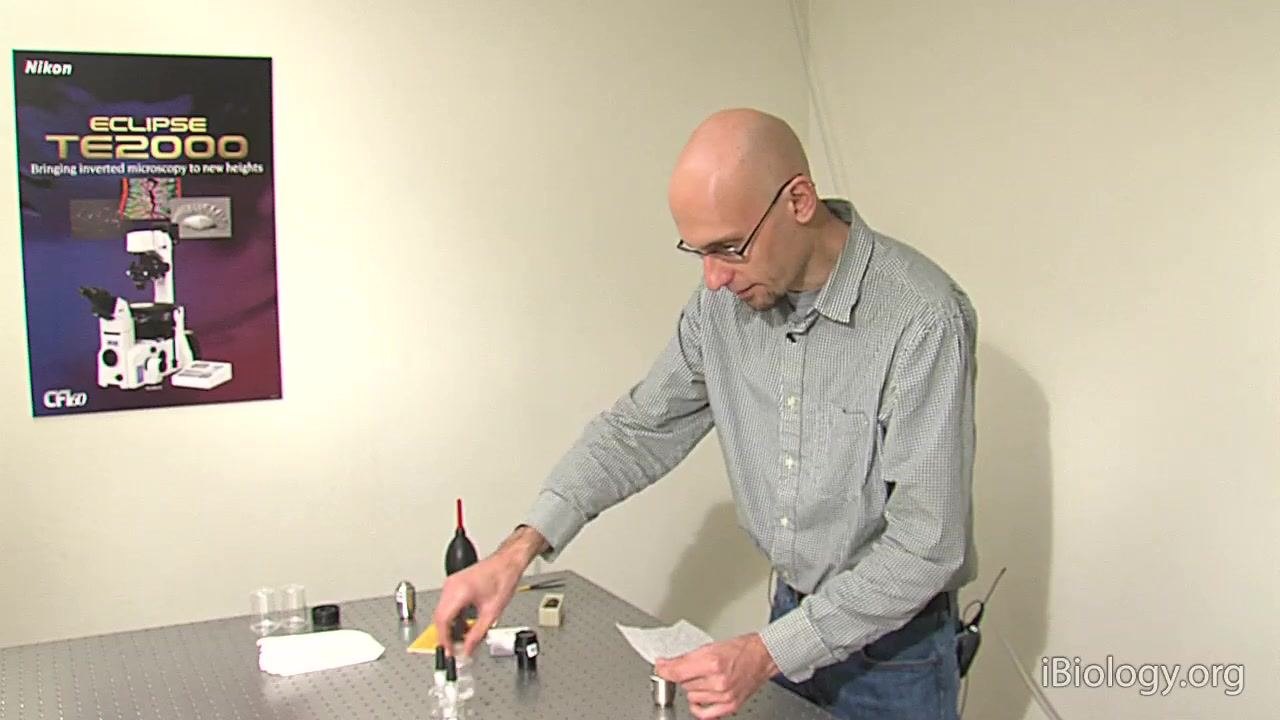 A person applies a drop of solvent to a microscope objective on a laboratory table, with cleaning supplies and lens paper visible.