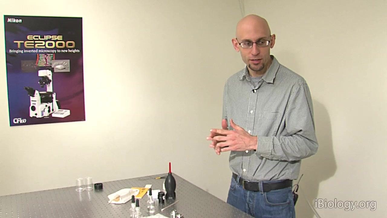 A person stands beside a table with microscope cleaning supplies, including bottles, a blower, and cloths, with a Nikon ECLIPSE TE2000 poster on the wall, emphasizing manufacturer-approved products.