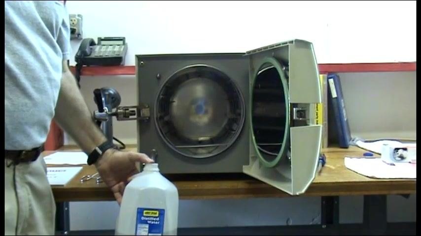The operator holding a labeled distilled water jug near the open autoclave chamber, preparing to refill the reservoir, with tools and paperwork nearby on the workbench.