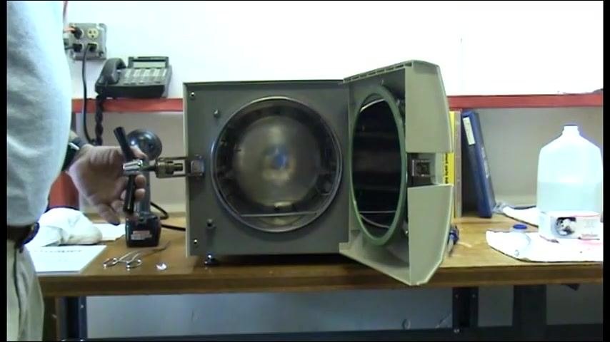 The autoclave with its door open, chamber exposed, and tools and cleaning supplies on the workbench as a person holds a tool near the unit.