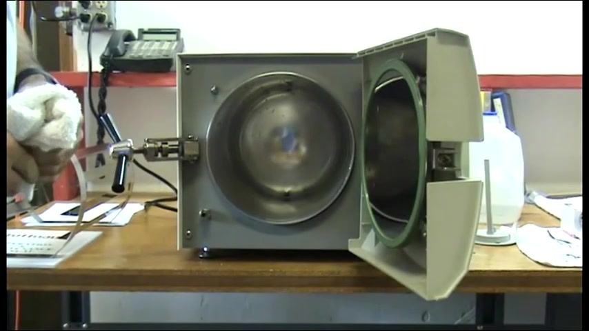 The autoclave chamber open with a person holding a white cloth nearby, control panel and supplies visible on the workbench.