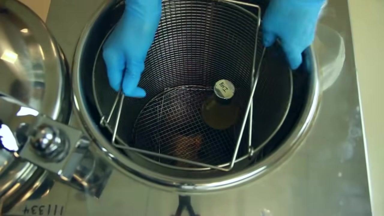 A person in a white lab coat and blue gloves stands next to a closed autoclave in a laboratory, preparing to operate it