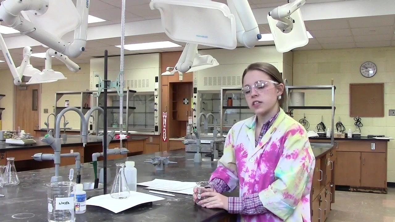 Laboratory workspace showing a burette, beakers, a waste container, and safety equipment, with a person wearing goggles and a lab coat.