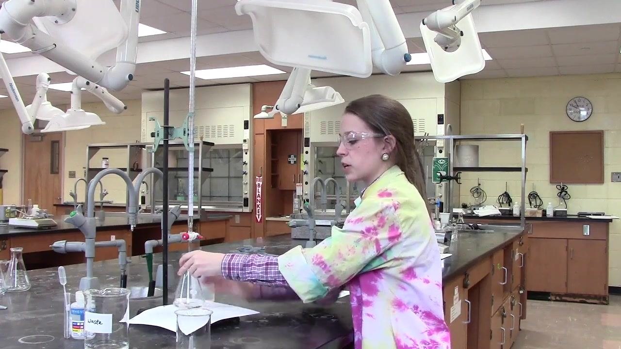 Person operating the burette with one hand while swirling the flask with the other, in a laboratory setting with labeled glassware and white paper beneath the flask.