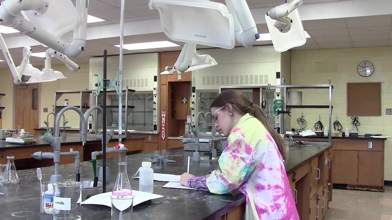 Student at a laboratory bench writing in a lab notebook or data sheet, with a burette, labeled glassware including a beaker labeled "Waste," a flask with a pink solution, a wash bottle, and other standard lab equipment visible in a well-equipped chemistry laboratory.