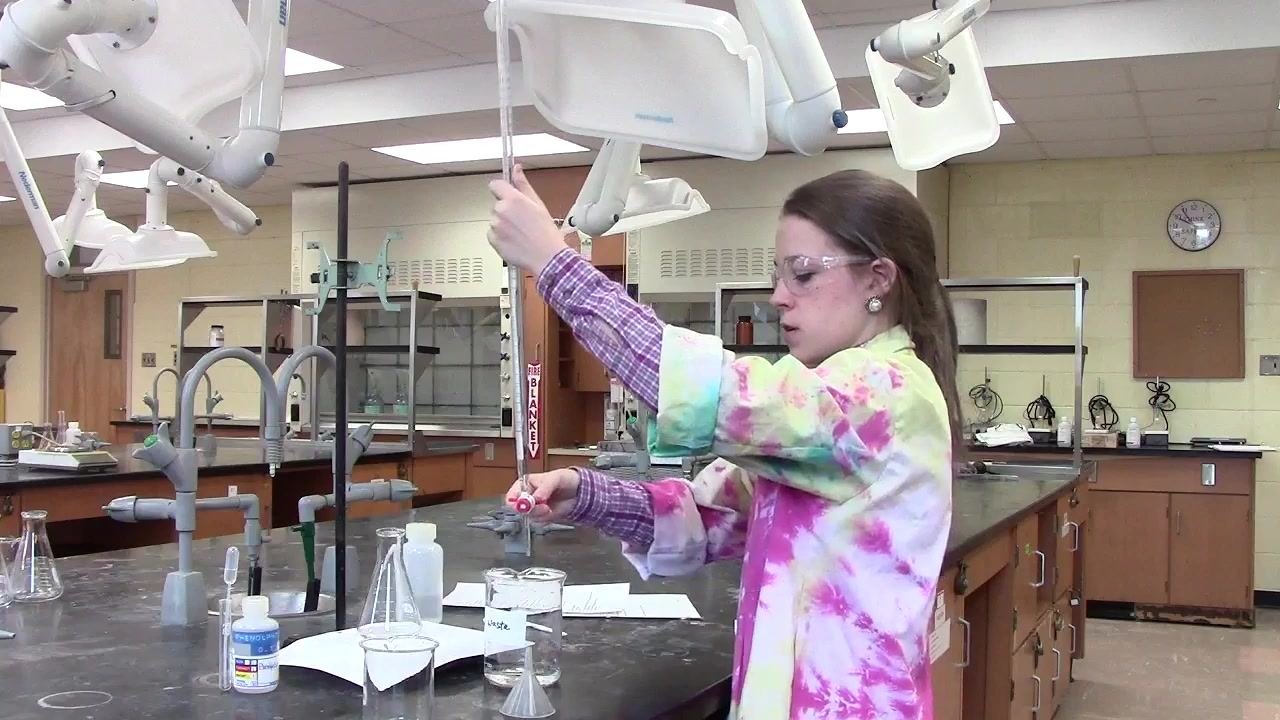 Person closing the burette stopcock in a laboratory setting, with a labeled waste beaker and other glassware visible.