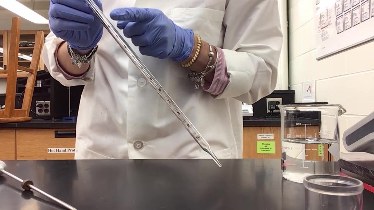 Lab technician in a lab coat and blue gloves holding a glass pipette vertically, aligning the meniscus with the zero mark, next to a beaker of water on the bench