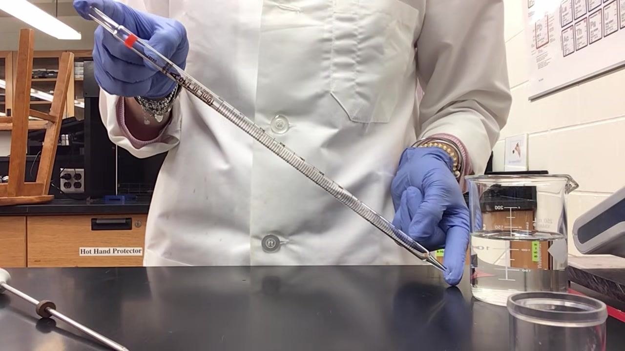 Lab technician in a lab coat and gloves holding the pipette while gesturing to explain calibration and measurement, with a beaker in the laboratory background