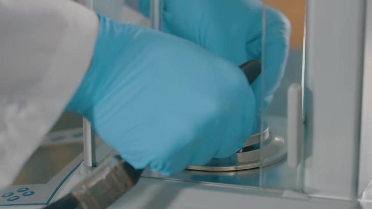 Gloved hands using a vacuum or cleaning tool inside the analytical balance weigh pan