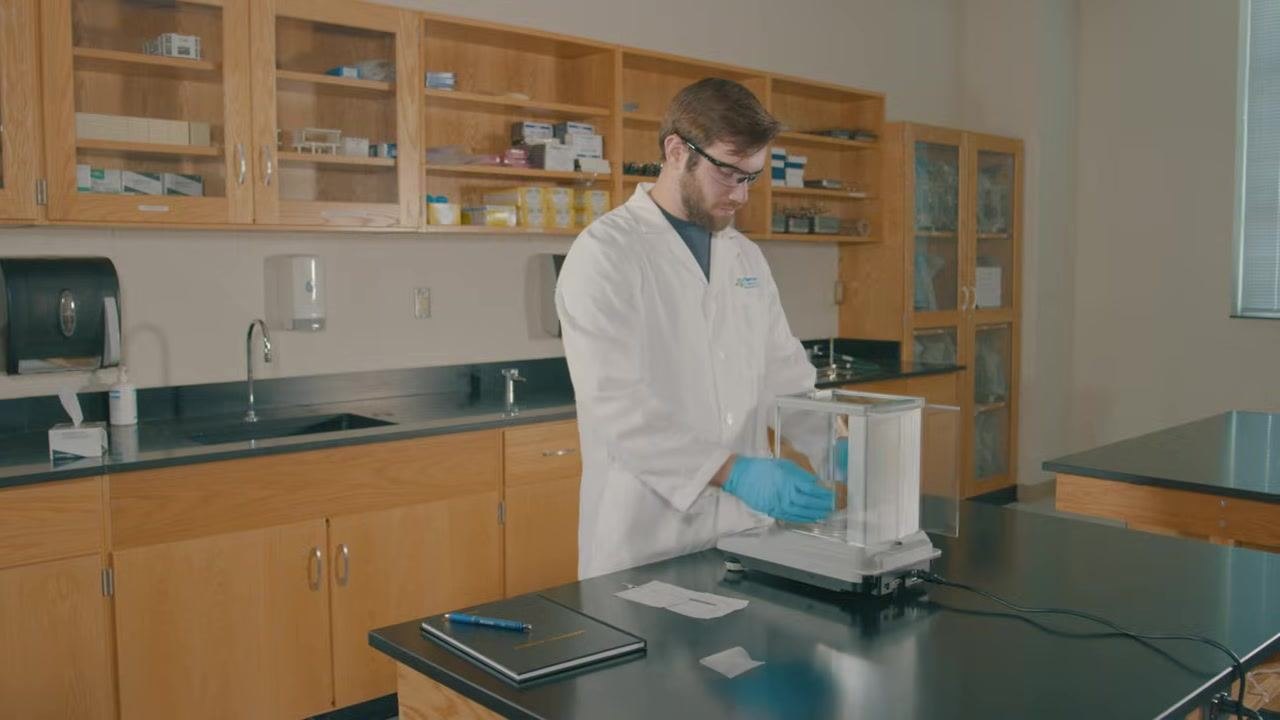 Lab technician in a lab coat and gloves closing the glass doors of the balance after adding sodium bicarbonate