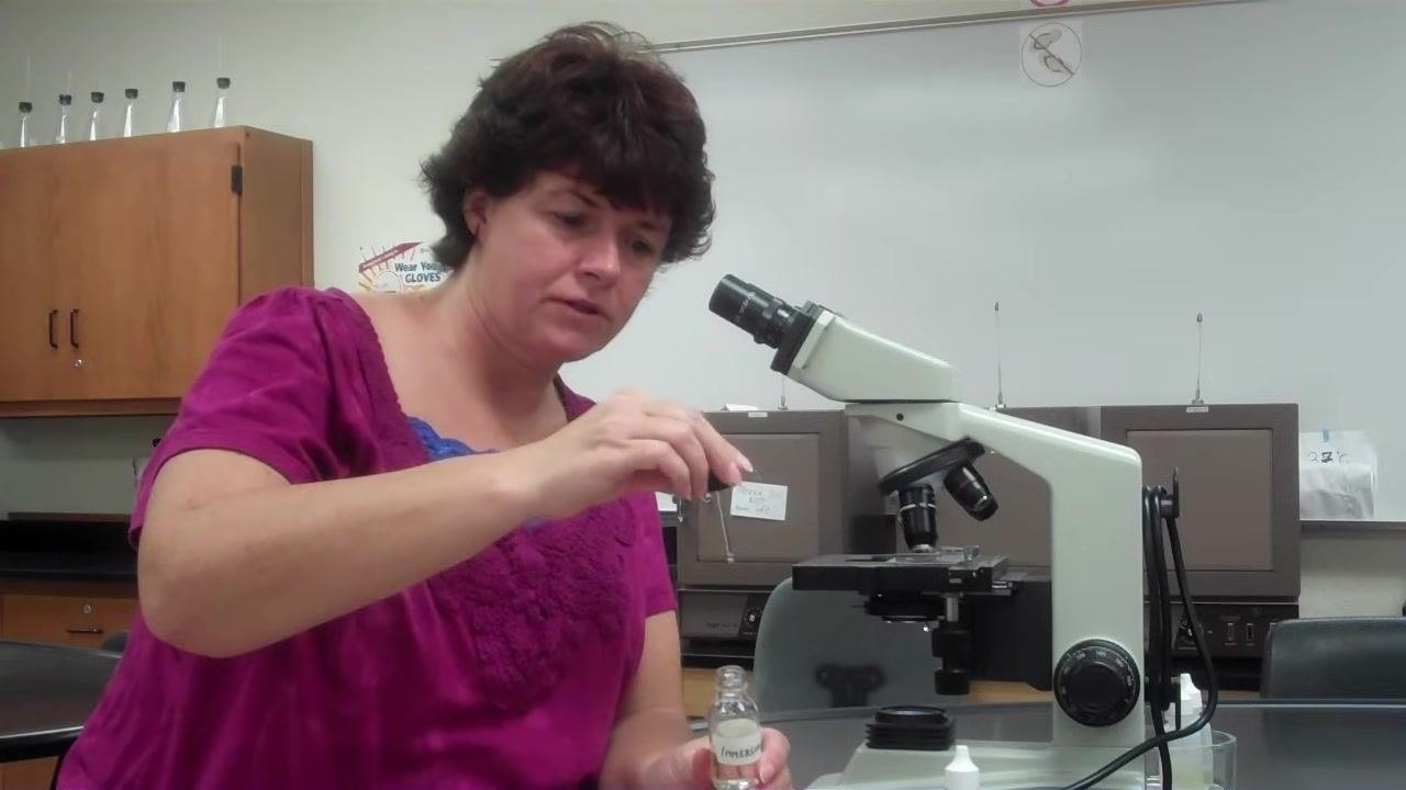 Instructor holding an immersion oil bottle and using a dropper to prepare a drop of oil above the slide, demonstrating proper technique.