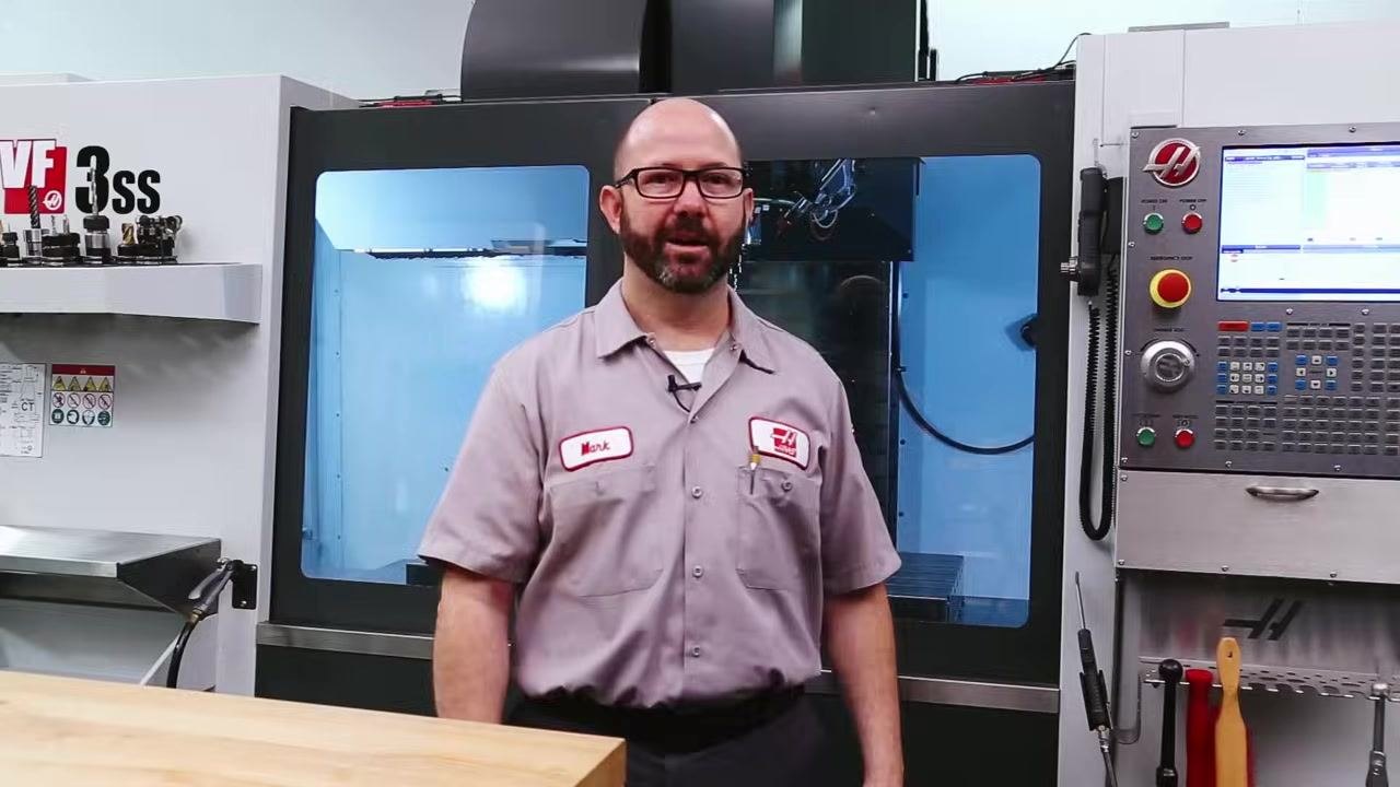 Whiteboard diagram showing a path from a house icon down to two labeled points, SPEED and FEED, drawn in a machine shop with CNC equipment and tool cabinets in the background.