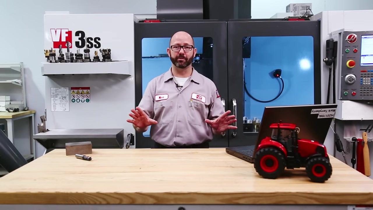 Workbench with a block of steel, cutting tool, laptop, and model tractor, set up in front of the CNC machine.