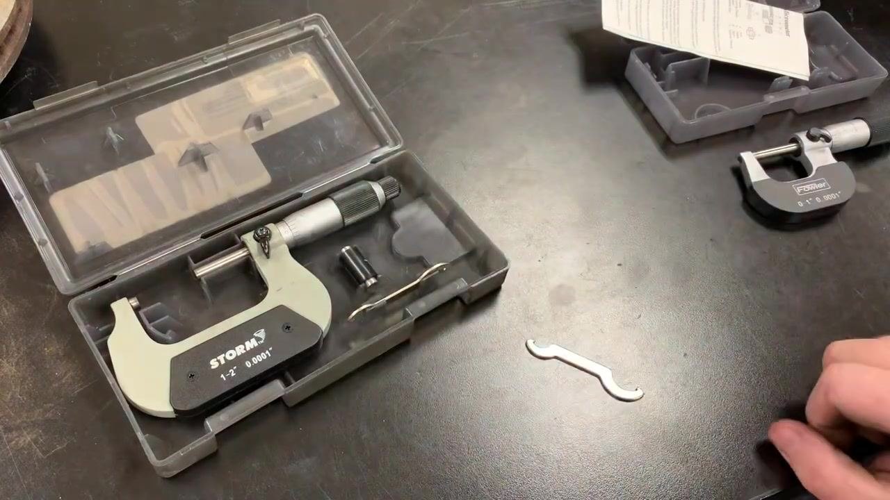 A STORM 1–2 inch micrometer in its case, with the reference standard and spanner wrench visible; a hand is reaching for the standard