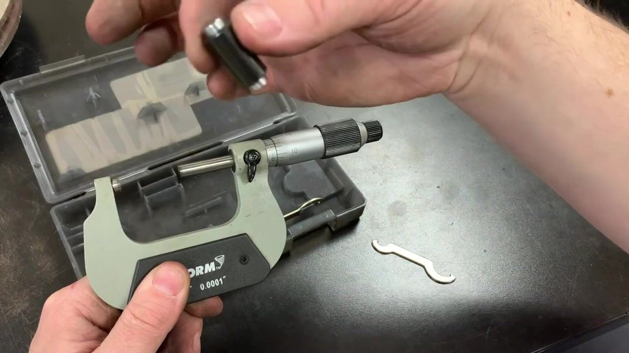 Hands holding a micrometer showing the zero marks perfectly aligned after calibration, with a workbench of micrometer cases and tools in the background