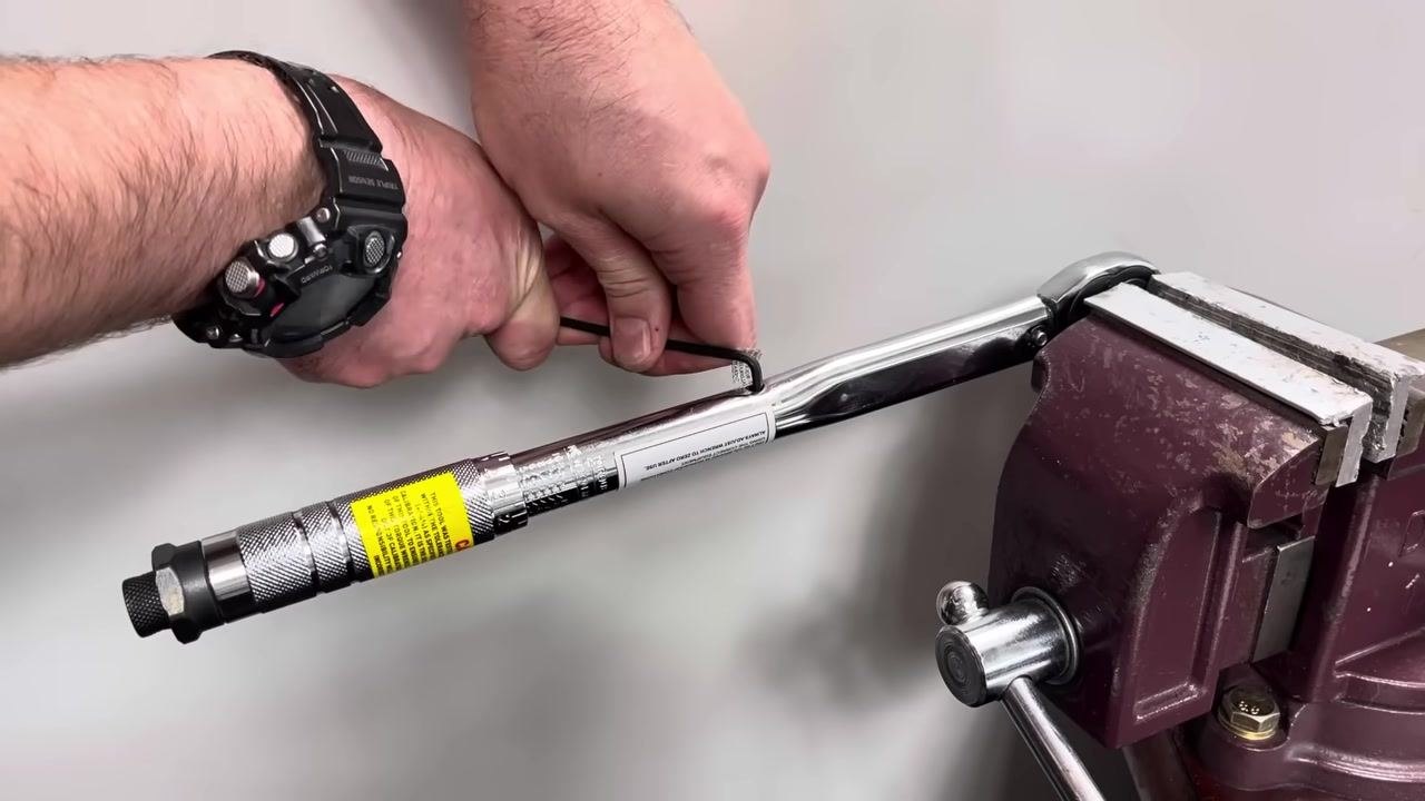 A hand uses an Allen key to adjust the calibration screw on the torque wrench while it is secured in the vise.
