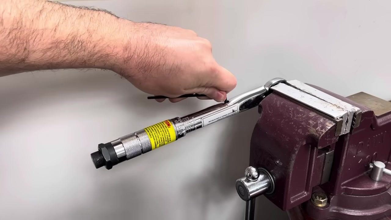 A hand adjusts the calibration screw on a torque wrench clamped in a vice in a workshop setting.