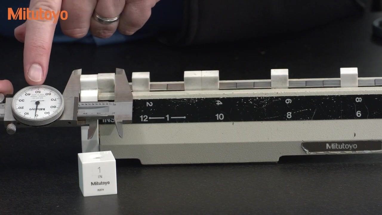 Close-up of a dial caliper measuring 1 inch on the calibration fixture, pointer at the 12 o'clock position, with a 1-inch Mitutoyo gauge block visible