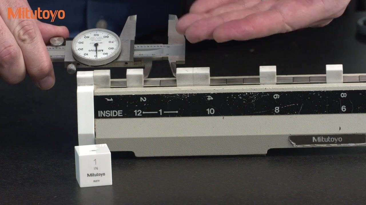 A dial caliper performing an inside, depth, or step measurement on the calibration fixture, pointer at the 12 o'clock position, with a 1-inch gauge block visible