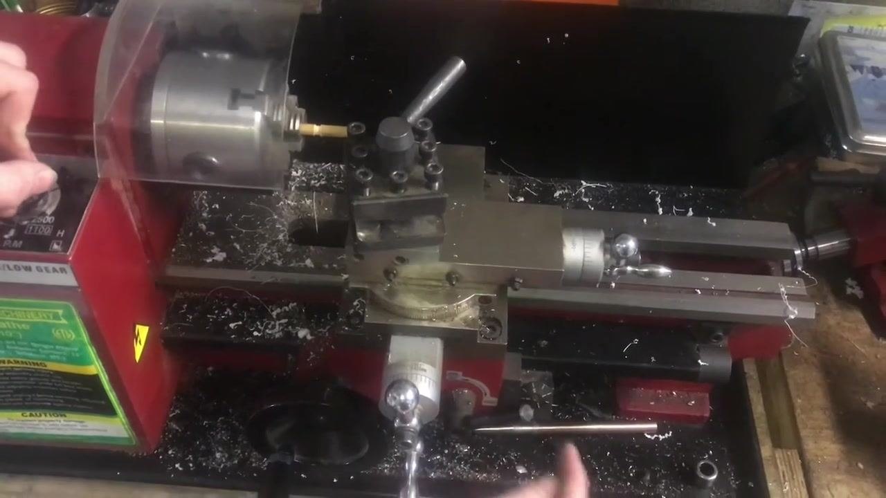 An overhead view of a mini lathe in operation. One hand is on the lathe control panel while the other is near the carriage handwheel and half-nut lever. The threading tool is set up in the tool post, metal shavings are visible on the lathe bed, the clear gear cover is in place, and the workpiece is chucked.