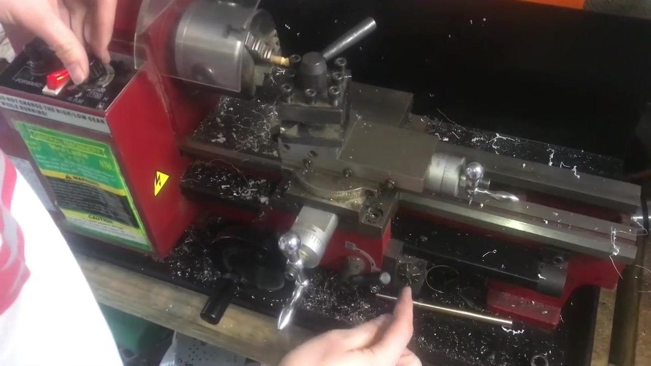 An operator's hand on the lathe control panel, showing the Forward/Reverse switch, RPM dial, and gear selector, with metal shavings in the work area and the tool post set up for threading.
