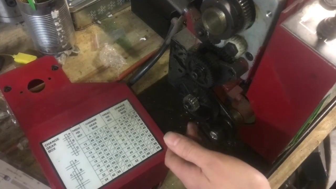 A hand pointing at and adjusting the gear train and banjo assembly, with the threading chart visible on the lathe cover and several gears exposed.