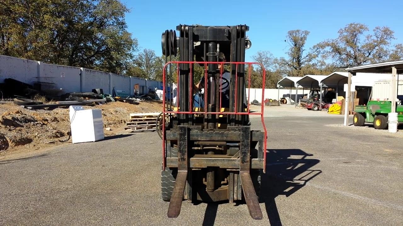 Operator's hand on the center hydraulic lever demonstrating tilt control, with the mast and hydraulic lines visible through the windshield.